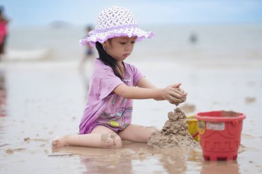 Balikpapan, 1 Mart 2023. Mor plaj şapkası ve tişörtü olan şirin küçük bir kız kumsalda kumsalda oynuyor. Tatil ve rahatlama. Yaz tatilinde sahilde aktif çocuk ve çocuk gelişimi..