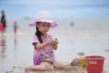Balikpapan, 1 Mart 2023. Mor plaj şapkası ve tişörtü olan şirin küçük bir kız kumsalda kumsalda oynuyor. Tatil ve rahatlama. Yaz tatilinde sahilde aktif çocuk ve çocuk gelişimi..