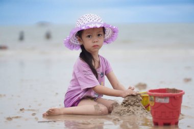 Balikpapan, 1 Mart 2023. Mor plaj şapkası ve tişörtü olan şirin küçük bir kız kumsalda kumsalda oynuyor. Tatil ve rahatlama. Yaz tatilinde sahilde aktif çocuk ve çocuk gelişimi..