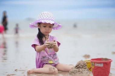 Balikpapan, 1 Mart 2023. Mor plaj şapkası ve tişörtü olan şirin küçük bir kız kumsalda kumsalda oynuyor. Tatil ve rahatlama. Yaz tatilinde sahilde aktif çocuk ve çocuk gelişimi..