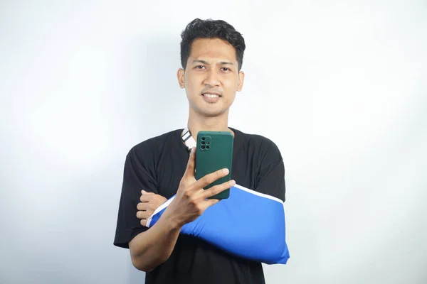 stock image Young man in casual clothes with broken hand using cellphone to talk to doctor via video conference