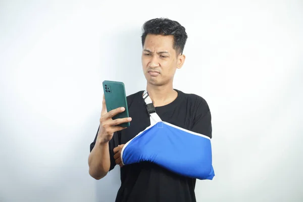 stock image Young man in casual clothes with broken hand using cellphone to talk to doctor via video conference