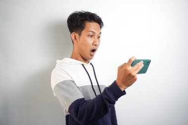 Cep telefonu ekranına bakan genç Asyalı şok olmuş bir adam çevrimiçi haberlere tepki veriyor. Ağzı açık ve beyaz arka planda izole edilmiş.