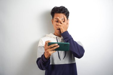 Cep telefonu ekranına bakan genç Asyalı şok olmuş bir adam çevrimiçi haberlere tepki veriyor. Ağzı açık ve beyaz arka planda izole edilmiş.