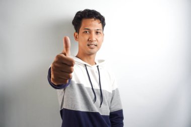 Kapüşonlu beyaz arka plan giyen yakışıklı bir Asyalı. Başparmakları havada. Çünkü iyi bir şey oldu.