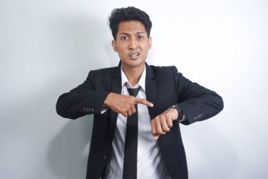 İzole edilmiş beyaz arka planda duran yakışıklı Asyalı genç adam saati izlemek için acele ediyor, sabırsız, sinirli ve geç kaldığı için kızgın.