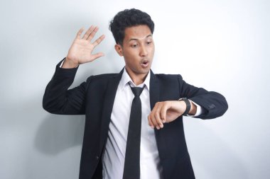 İzole edilmiş beyaz arka planda duran yakışıklı Asyalı genç adam saati izlemek için acele ediyor, sabırsız, sinirli ve geç kaldığı için kızgın.