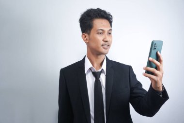 Beyaz arka planda yalnız duran, cep telefonu kullanan, neşeli, heyecanlı, takım elbiseli Asyalı genç bir adamın portresi.