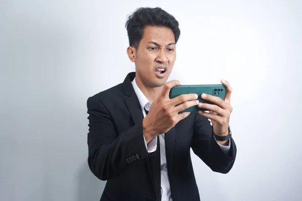 愤怒的亚洲年轻人在工作中玩游戏时 会在智能手机上生气 身穿西服的印度尼西亚男子 — 图库照片