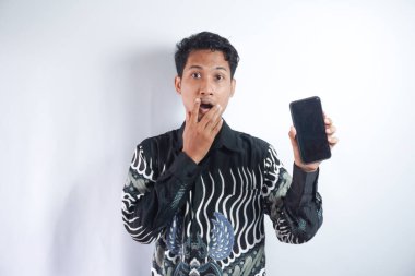 Gri arka planda izole edilmiş Batik tişörtlü yakışıklı bir adam fotoğrafı. Akıllı telefon sunuyor ve reklam için fotokopi çekilen boş siyah ekranı işaret ediyor. Şaşkınlık ve haz ifadesi