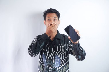 Gri arka planda izole edilmiş Batik tişörtlü yakışıklı bir adam fotoğrafı. Akıllı telefon sunuyor ve reklam için fotokopi çekilen boş siyah ekranı işaret ediyor. Şaşkınlık ve haz ifadesi