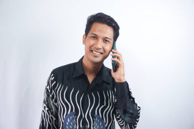 Genç, neşeli, mutlu, 20 'li yaşlarda beyaz bir adam Batik tişört giyiyor cep telefonuyla konuşuyor ve beyaz arka plan stüdyo fotoğraflarında izole edilmiş bir şekilde gülümsüyor.