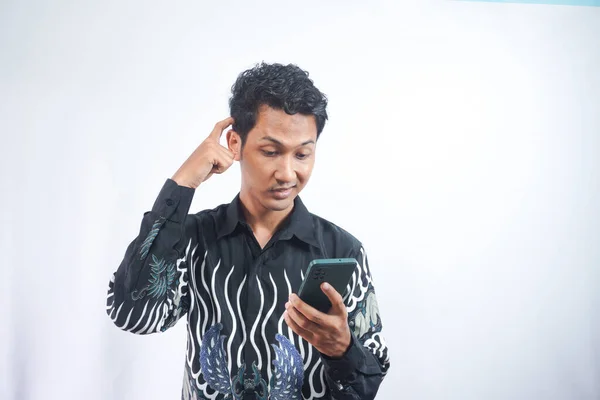 Portrait of handsome young adult with dreamy look, thinking while holding smartphone, isolated over white background.