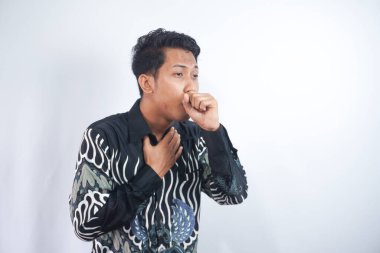 Batik gömlekli bir adam yumruğuna öksürüyor, gri bir arka planda izole edilmiş.