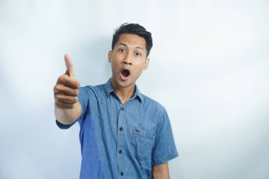 Asyalı yakışıklı, mavi gömlekli, soyutlanmış beyaz arka planı olan genç bir adam. Başparmakları havada çünkü iyi bir şey oldu.