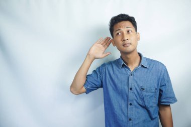 Genç Asyalı çılgın adam ciddi ve meraklı görünüyor, dinliyor, gizli bir konuşma ya da dedikodu duymaya çalışıyor, düz renkli duvara kulak misafiri oluyor.