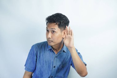 Genç Asyalı çılgın adam ciddi ve meraklı görünüyor, dinliyor, gizli bir konuşma ya da dedikodu duymaya çalışıyor, düz renkli duvara kulak misafiri oluyor.