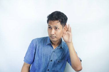 Genç Asyalı çılgın adam ciddi ve meraklı görünüyor, dinliyor, gizli bir konuşma ya da dedikodu duymaya çalışıyor, düz renkli duvara kulak misafiri oluyor.