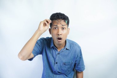 Şaşkın Asyalı genç adam gözlüklerini çıkar, şaşkın ve hoşnutsuz bir yüz ifadesiyle bak, garip ve korkunç bir şeyden kork, mavi gömleğin içinde beyaz arka planda dur.