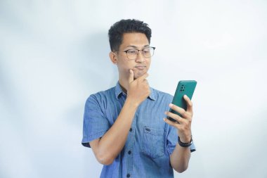 Mavi gömlekli Asyalı bir adam telefonuna ciddi ciddi bakıyor ve stüdyoda beyaz bir arka planda gözlük takıyor..