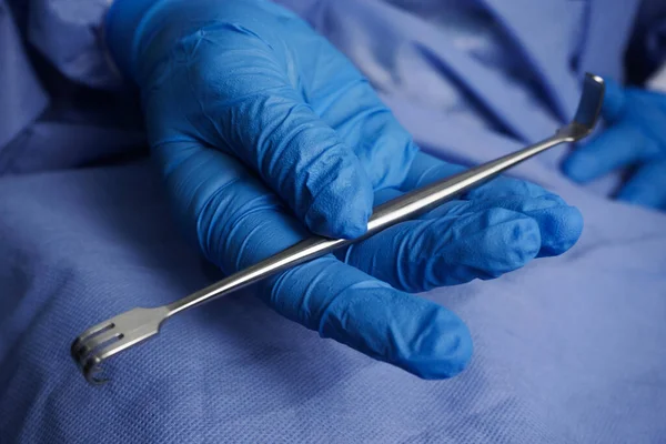 Keskin Senn-Miller Retraktörü tutan mavi eldivenli doktor çok amaçlı, çift uçlu bir çekiş cihazıdır. Genel cerrahi işlemlerde yüzeysel yaraları açmak için kullanılabilir.