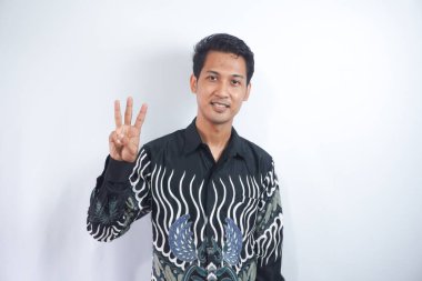 Soyutlanmış arka planda genç yakışıklı bir adam üç numarayı gösterip işaret ederken kendinden emin ve mutlu bir şekilde gülümsüyor..