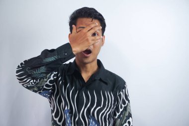 Şok olmuş Batik gömlekli genç iş adamı beyaz arka planda poz verirken elleriyle gözlerini kapattı.