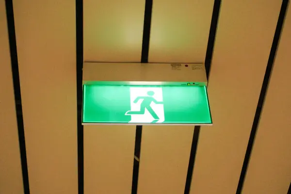 A green fire exit sign is placed on the ceiling along the dimly lit corridor and there is green exit sign on the exit door.