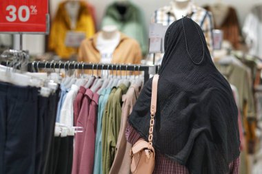 Alışveriş merkezindeki giyim ürünlerine bakan tesettürlü genç, güzel Asyalı bir kadın. Çekici, mutlu kız alışveriş merkezindeki elbise raflarından yeni kıyafetler ve tişörtler seçmek için mağazada gezinmekten zevk alıyor..