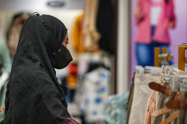 Alışveriş merkezindeki giyim ürünlerine bakan tesettürlü genç, güzel Asyalı bir kadın. Çekici, mutlu kız alışveriş merkezindeki elbise raflarından yeni kıyafetler ve tişörtler seçmek için mağazada gezinmekten zevk alıyor..