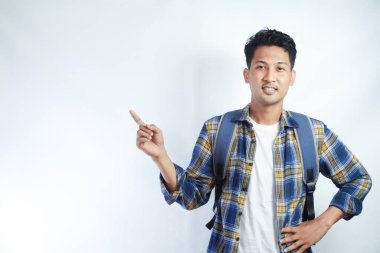 Neşeli Asyalı erkek öğrenci günlük kıyafetler ve sırt çantası gözlükleriyle beyaz arka planda izole edilmiş boş alanı işaret ediyor. Lise üniversite konsepti