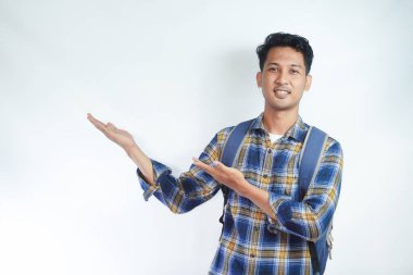 Neşeli Asyalı erkek öğrenci günlük kıyafetler ve sırt çantası gözlükleriyle beyaz arka planda izole edilmiş boş alanı işaret ediyor. Lise üniversite konsepti