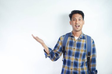 Neşeli Asyalı erkek öğrenci günlük kıyafetler ve sırt çantası gözlükleriyle beyaz arka planda izole edilmiş boş alanı işaret ediyor. Lise üniversite konsepti