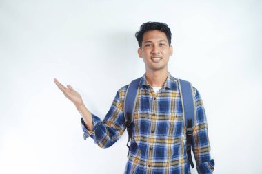 Neşeli Asyalı erkek öğrenci günlük kıyafetler ve sırt çantası gözlükleriyle beyaz arka planda izole edilmiş boş alanı işaret ediyor. Lise üniversite konsepti