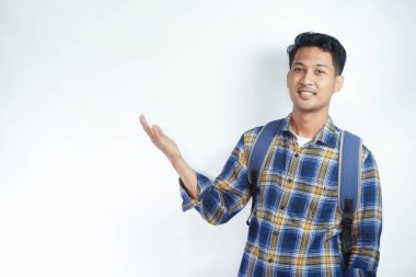 Neşeli Asyalı erkek öğrenci günlük kıyafetler ve sırt çantası gözlükleriyle beyaz arka planda izole edilmiş boş alanı işaret ediyor. Lise üniversite konsepti