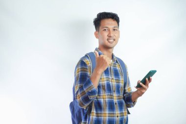 Sırt çantalı neşeli Asyalı genç bir öğrenci cep telefonunu tutarken ayakta duruyor ve beyaz arka planda izole bir şekilde baş parmağını gösteriyor. Lise üniversite konsepti