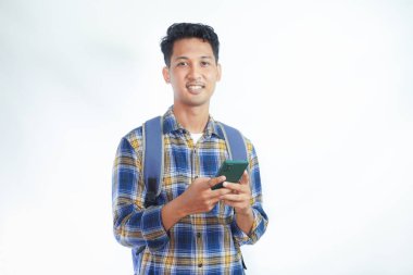 Sırt çantasıyla gülümseyen Asyalı genç bir öğrenci beyaz arka planda cep telefonunu elinde tutarken ayakta duruyor. Lise üniversite konsepti