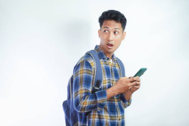 Sırt çantası gözlüklü genç Asyalı şoka uğramış bir öğrenci cep telefonunu elinde tutarken ve beyaz arka planda izole edilmiş ağzını açarken ayakta duruyor. Lise üniversite konsepti