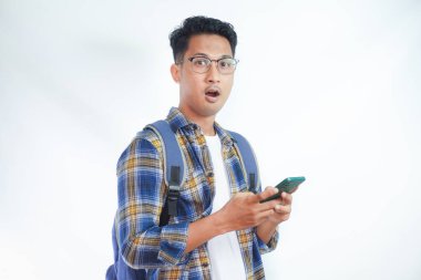 Sırt çantası gözlüklü genç Asyalı şoka uğramış bir öğrenci cep telefonunu elinde tutarken ve beyaz arka planda izole edilmiş ağzını açarken ayakta duruyor. Lise üniversite konsepti