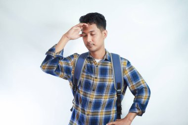 Sırt çantalı, yorgun, Asyalı genç bir öğrenci gözlerini kapalı, elini başının üstüne koyup beyaz arka planda izole bir şekilde duruyor. Lise üniversite konsepti