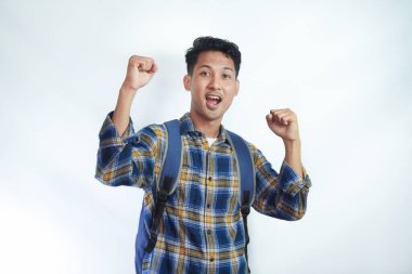 Sırt çantalı flanel gömlek giyen heyecanlı Asyalı genç erkek öğrenci kazanan jestler yapıyor ve beyaz arka planda izole edilmiş başarıyı kutluyor. Lise üniversite konsepti eğitimi