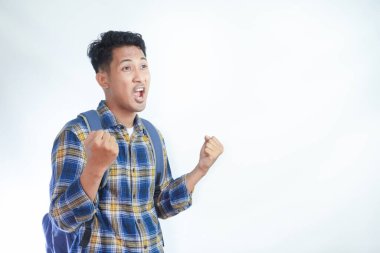 Sırt çantalı flanel gömlek giyen heyecanlı Asyalı genç erkek öğrenci kazanan jestler yapıyor ve beyaz arka planda izole edilmiş başarıyı kutluyor. Lise üniversite konsepti eğitimi