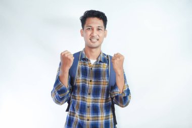 Sırt çantalı flanel gömlek giyen heyecanlı Asyalı genç erkek öğrenci kazanan jestler yapıyor ve beyaz arka planda izole edilmiş başarıyı kutluyor. Lise üniversite konsepti eğitimi