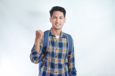 Sırt çantalı flanel gömlek giyen heyecanlı Asyalı genç erkek öğrenci kazanan jestler yapıyor ve beyaz arka planda izole edilmiş başarıyı kutluyor. Lise üniversite konsepti eğitimi