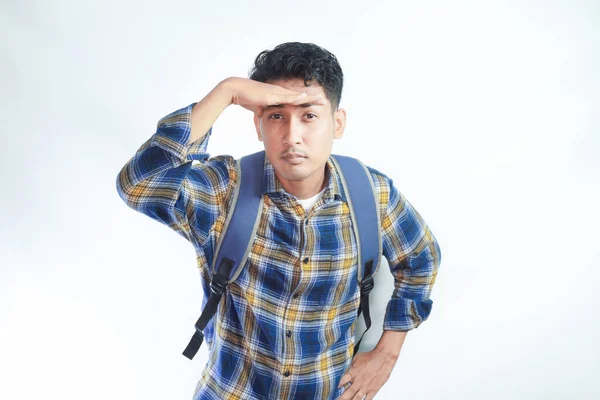 serious young Asian student man in casual clothes with backpack hold hand at his forehead looking at the camera isolated on white background. High school university college concept