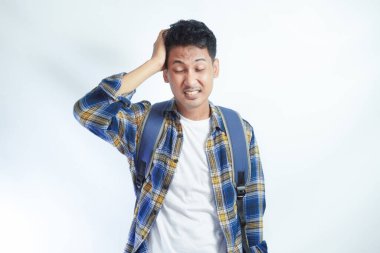 Sırt çantalı, yorgun, Asyalı genç bir öğrenci gözlerini kapalı, elini başının üstüne koyup beyaz arka planda izole bir şekilde duruyor. Lise üniversite konsepti