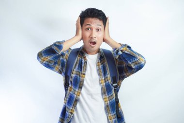 Ciddi ve şok olmuş genç Asyalı adam, günlük gömlekli, kulaklarının yanında el ele tutuşmuş ve ağzı açık, beyaz arka planda izole edilmiş dedikoduları dinliyor.