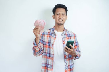 Adult Asian man holding money and mobile phone smiling happy at the camera clipart