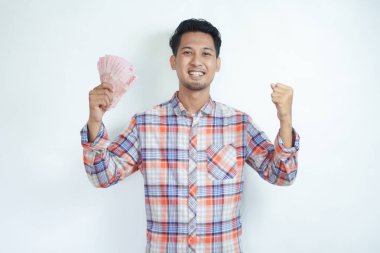 Adult Asian man clenched fist while holding paper money and showing happy expression clipart