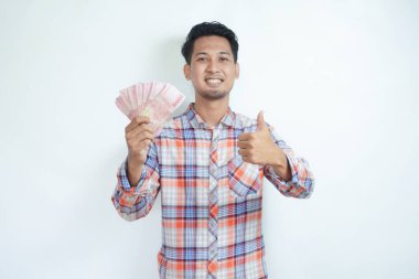 Yetişkin Asyalı bir adamın yakın plan portresi elinde para tutarken başparmağını kaldırır ve mutlu bir yüz ifadesi gösterir.